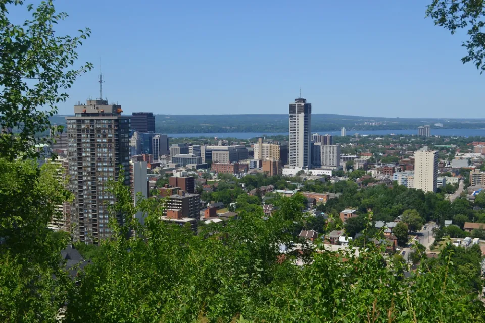 The downtown high rises of Hamilton, Ontario are surrounded by greenery and the blue of Lake Ontario on a sunny day.